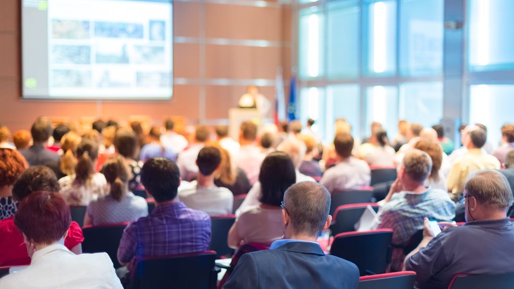 Men and Women Sitting at a Conference with Big Screen in Background
