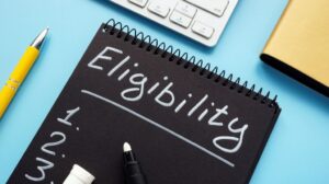 Black Notebook with White Eligibility Text and List on a Desk with Laptop